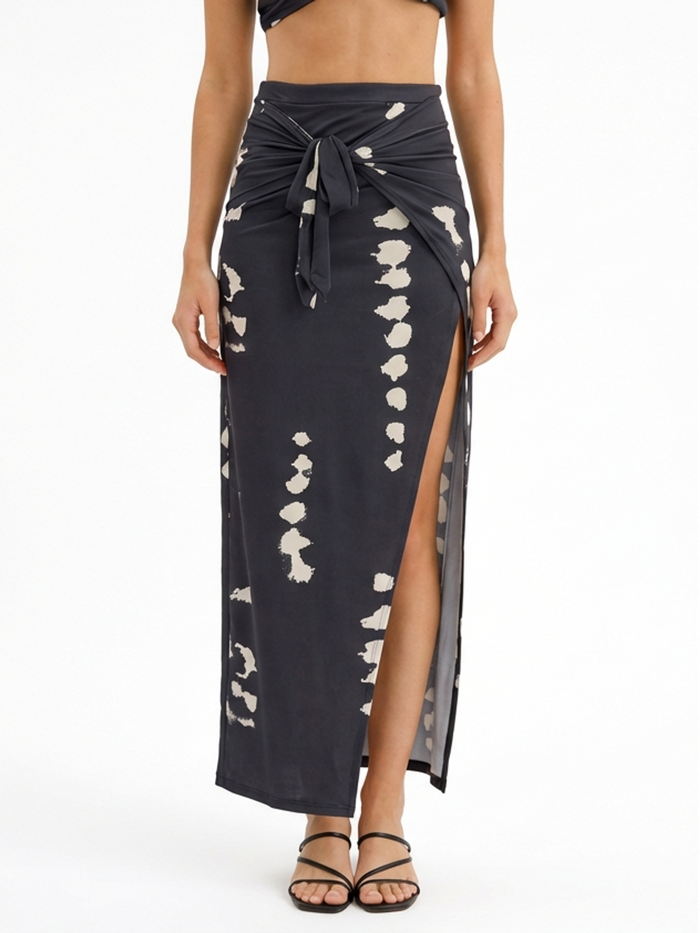 A woman wears a dark maxi skirt with a side slit and a knotted waist, featuring a light abstract pattern.