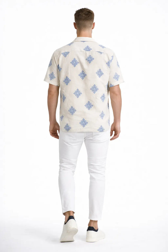 A man wearing a short-sleeve, patterned shirt with blue designs, paired with white pants and casual shoes.