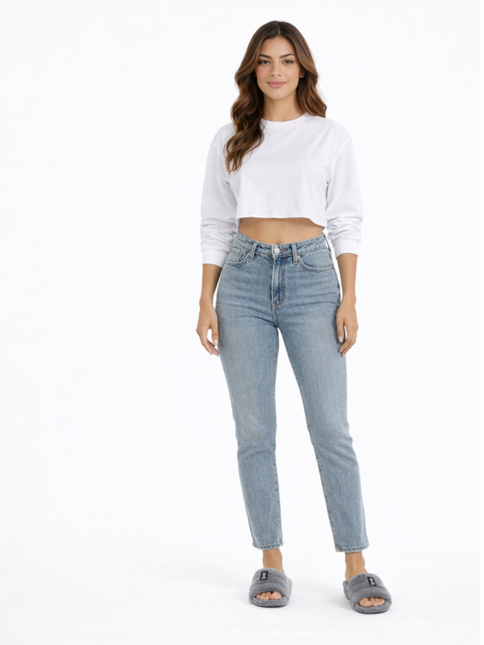 A woman stands confidently in a cropped white long-sleeve shirt and light blue jeans, paired with gray slippers.