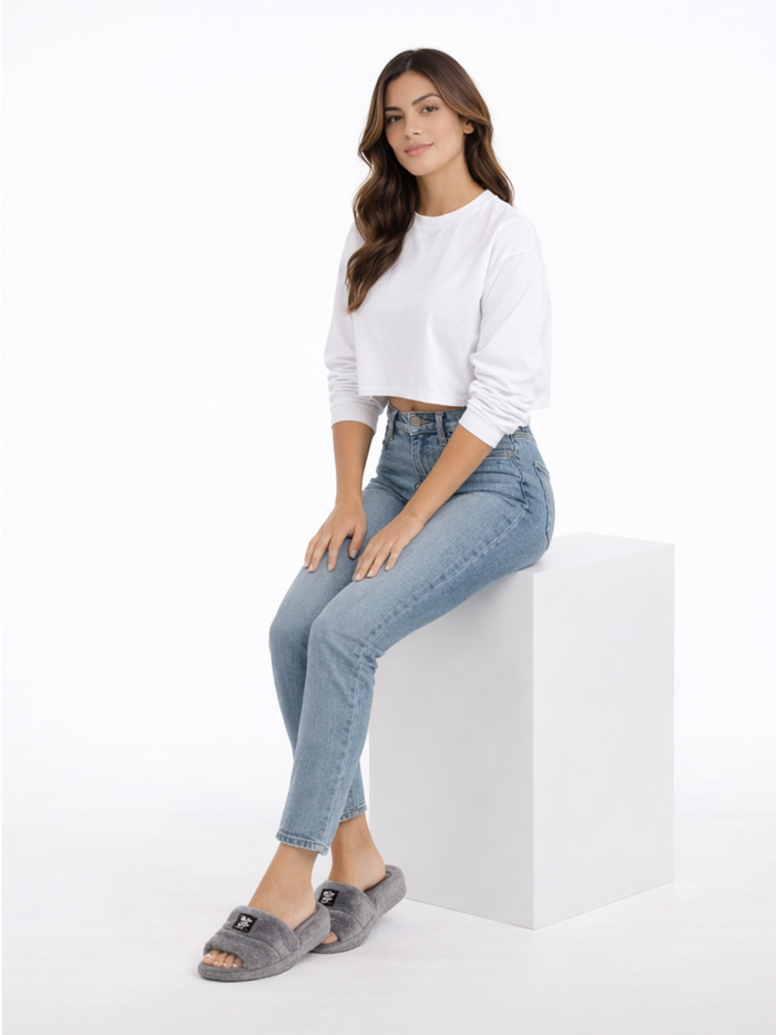 A woman sits on a white pedestal wearing a cropped white top, light blue jeans, and gray fuzzy slippers.