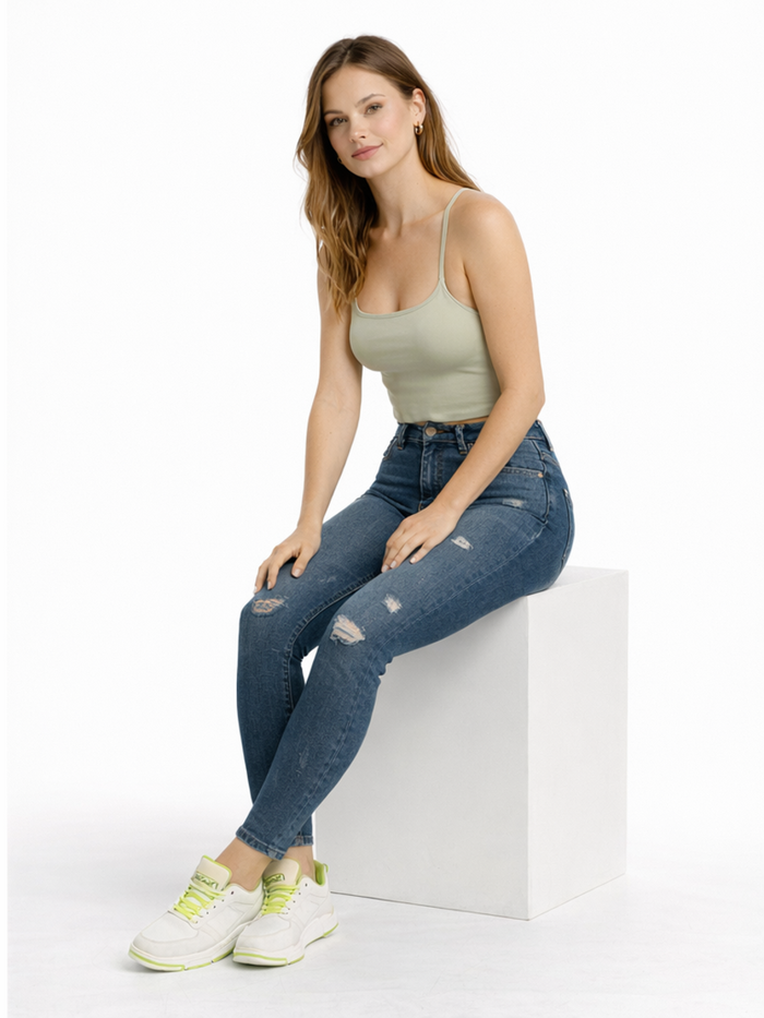 A woman sits on a white cube, wearing a light green tank top, distressed blue jeans, and white sneakers with neon acc...