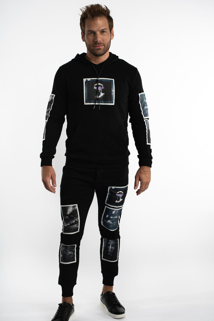 Model in black distressed joggers with blind frame graphic patches, styled with a matching hoodie and black sneakers.