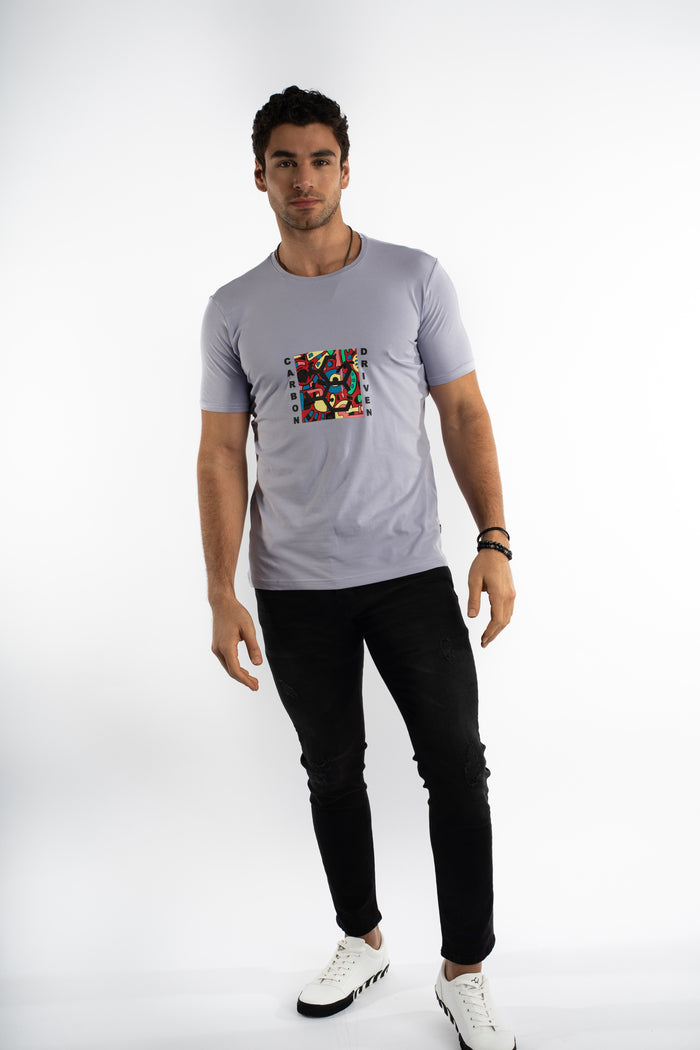 Man in lilac Carbon Driven Graphic Tee with colorful chest art, black distressed jeans, and white sneakers.