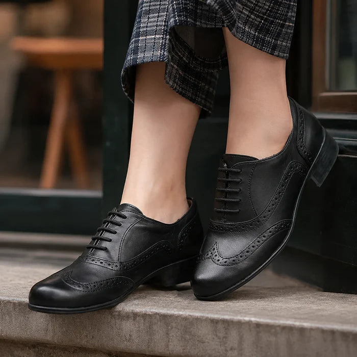 Black leather brogue lace-up shoes with wingtip perforations, worn with pants