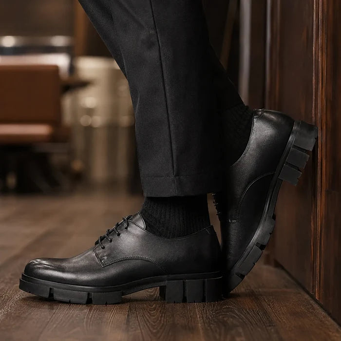 Black leather lace-up shoe with strap detail and chunky lug sole on wooden floor.