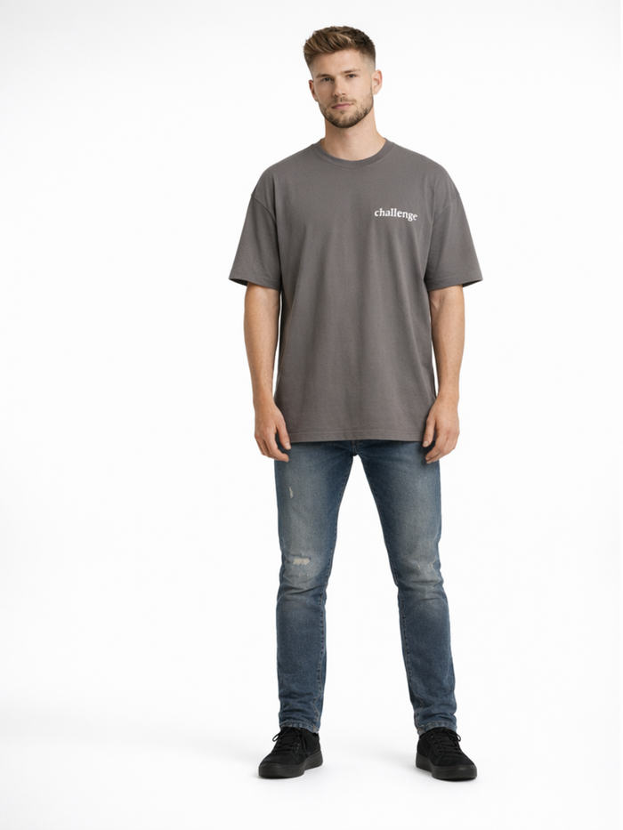 A man stands wearing a gray oversized t-shirt and distressed blue jeans, paired with black sneakers.