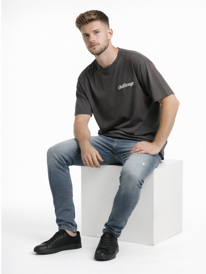 A man sits on a white cube, wearing a dark gray t-shirt with the word "challenge" and blue jeans, paired with black s...
