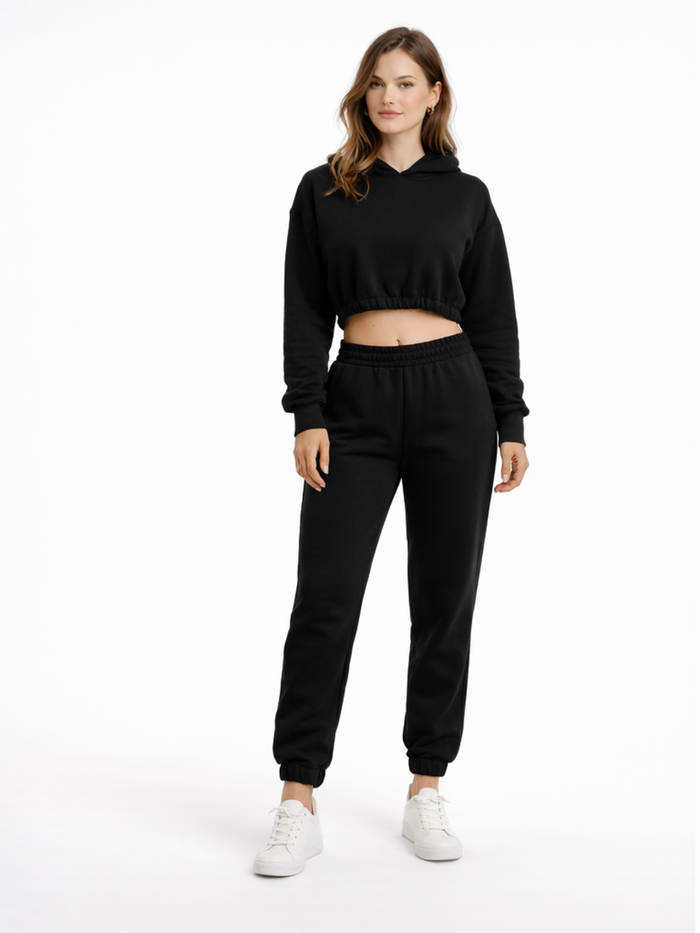 A model wearing a black cropped hoodie and matching joggers, paired with white sneakers, stands against a plain backg...