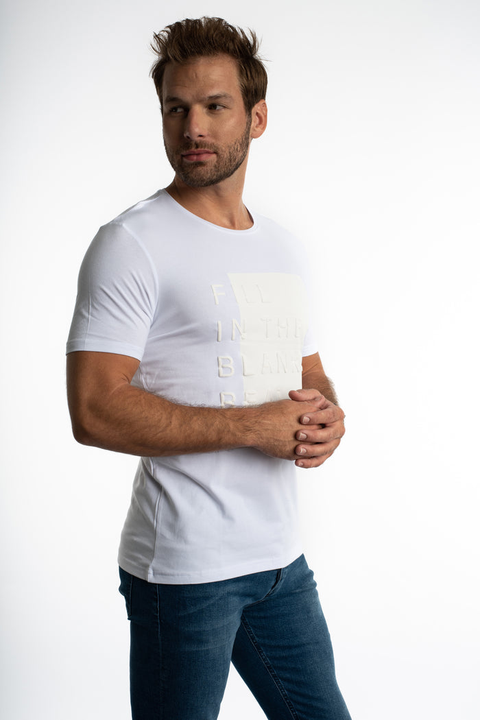 Man in a white Fill In The Blank streetwear t-shirt with an embossed graphic paired with blue denim jeans.