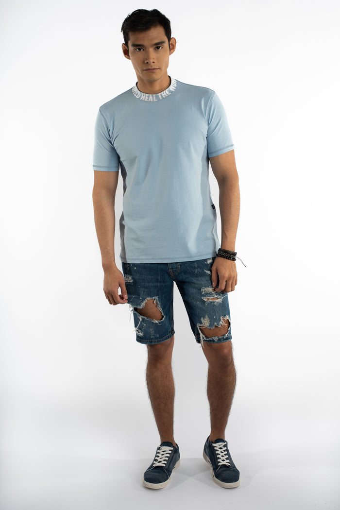 Light blue Heal The World graphic tee with embroidered collar text, worn by male model in ripped denim shorts.
