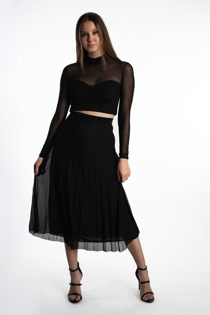 Model in Jada black high-waisted pleated midi skirt with mesh overlay, paired with a long-sleeve crop top and heels.