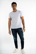 A man stands confidently in a fitted white t-shirt and distressed dark jeans, paired with white sneakers.