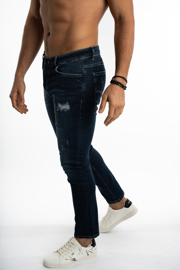 A man wearing dark blue distressed jeans and white sneakers poses against a plain background.