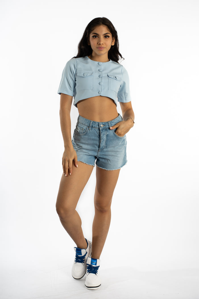 Model wearing Journi high-waisted blue washed denim shorts with raw hem and light blue cropped button-up top.