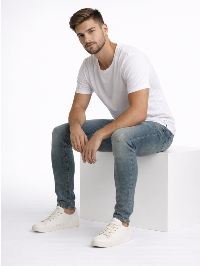 A man sits on a white cube, wearing a plain white t-shirt, light blue jeans, and casual white sneakers.