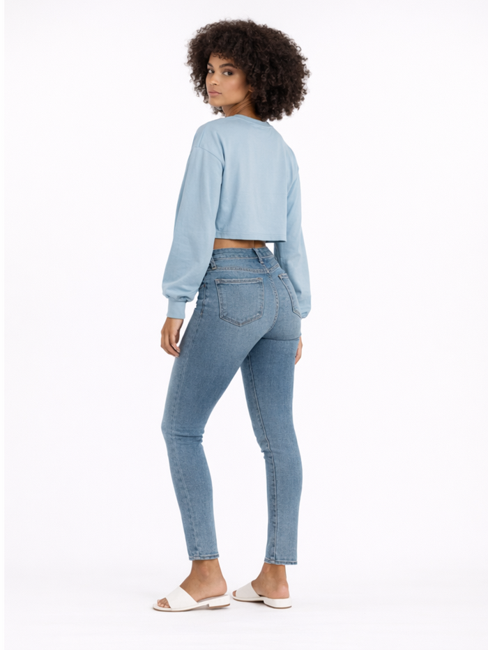 A model with curly hair wears a light blue cropped top, fitted jeans, and white sandals, posing from the back.