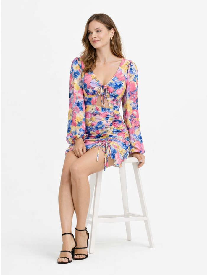 A model sits on a stool wearing a colorful floral dress with long sleeves and a cinched waist.