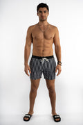 Man in Niko black and white striped swim trunks with white drawstring, Carbon brand slides, and poolside summer style.