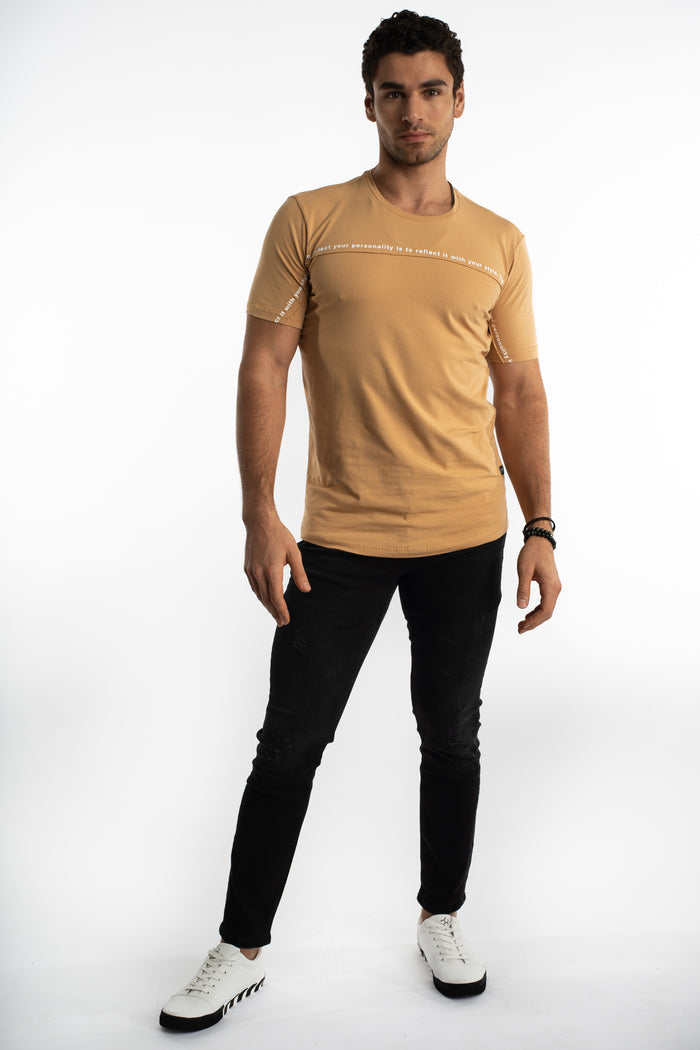 Man in Reflect Your Style tan graphic tee, black streetwear jeans, and white sneakers against white background.