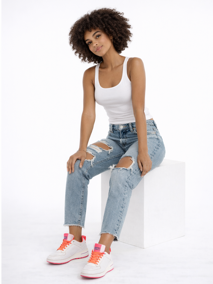 A model poses on a white block, wearing a fitted tank top, distressed jeans, and colorful sneakers.