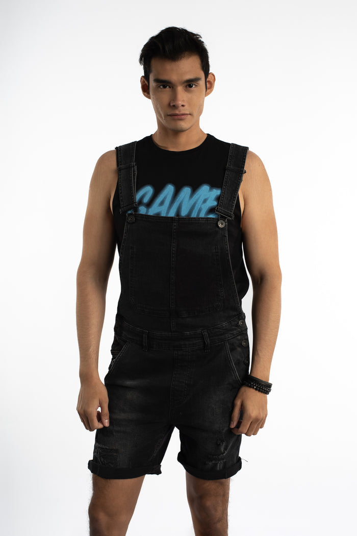 Man wearing Wayne Black Denim Overalls in carbon with distressed shorts and adjustable straps over a tank top.