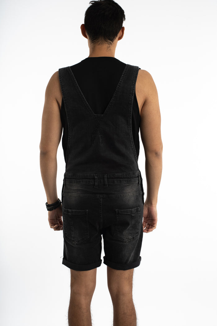 Back view of man in Wayne black denim overall shorts with V-shaped straps and a streetwear fit against white.