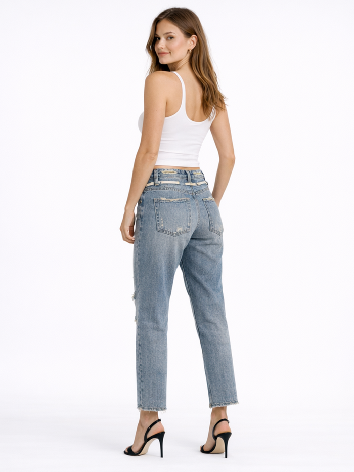 A woman stands with her back to the camera, wearing light blue jeans and a white tank top, paired with black heels.
